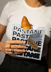 Person holding a branded paper bag filled with a croissant, wearing gold rings and a white top