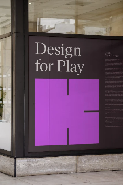 A purple graphic with text that reads, "Design for Play" mounted in a dark metal frame on a wall outside in a public setting.