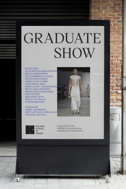Poster for a graduate show displaying students names and a runway image, set against a brick wall and metal siding outdoors.