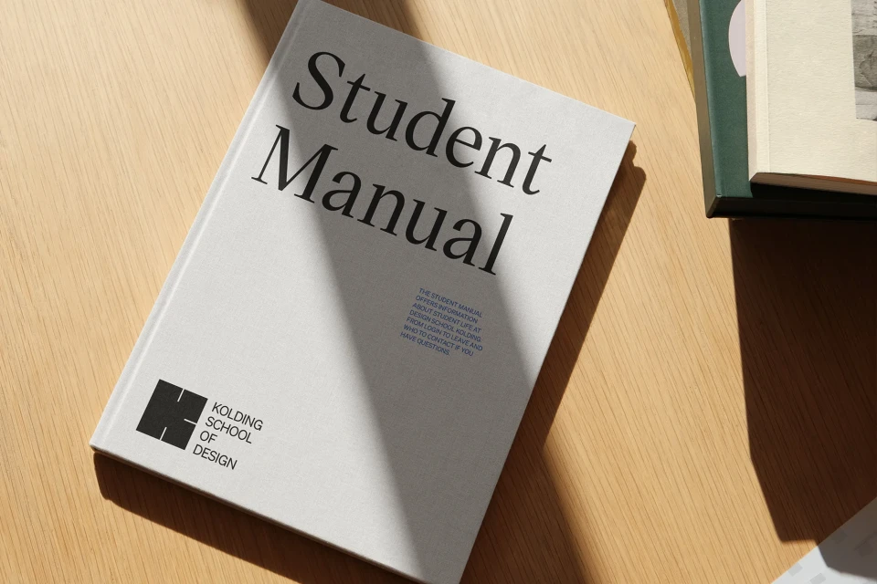 Student manual for Kolding School of Design on a wooden table with sunlight and shadows. Study aid, design education reference.