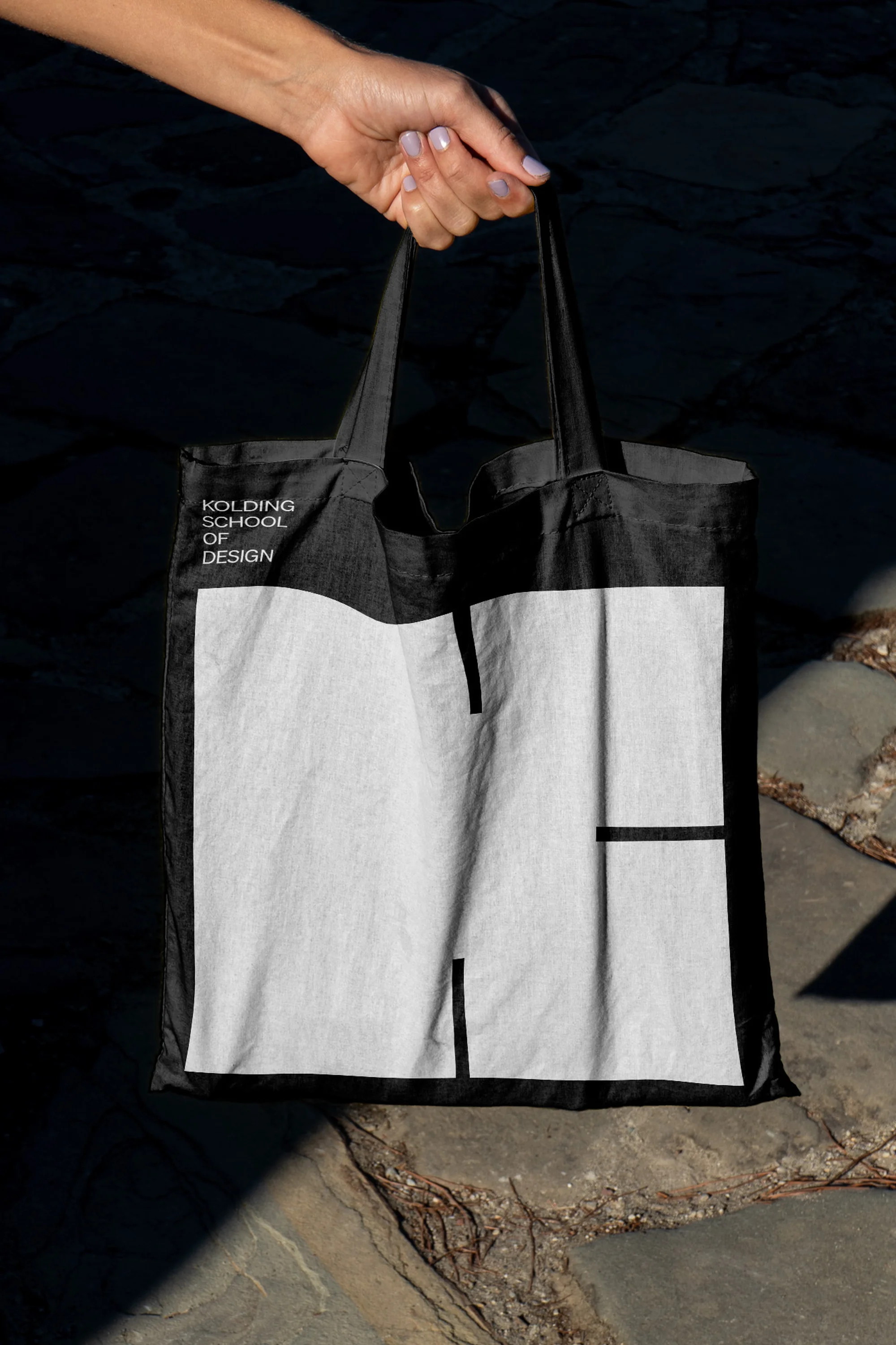 Black tote bag with white graphic logo for Kolding School of Design, held by a hand against a dark stone background.
