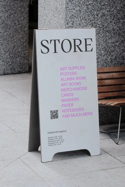 White A-frame sign displaying art supplies, posters, books, and merchandise. The sign stands on a tile surface outside.