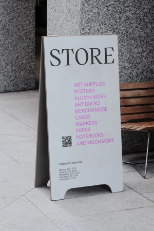 White A-frame sign displaying art supplies, posters, books, and merchandise. The sign stands on a tile surface outside.