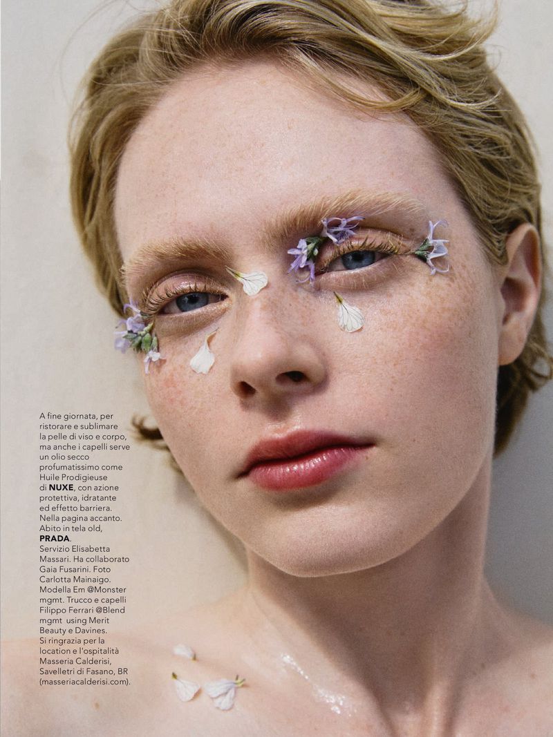 Close-up portrait of a fair-skinned young woman with blonde hair, featuring flowers around her eyes, enhancing her beauty in a minimalistic setting.