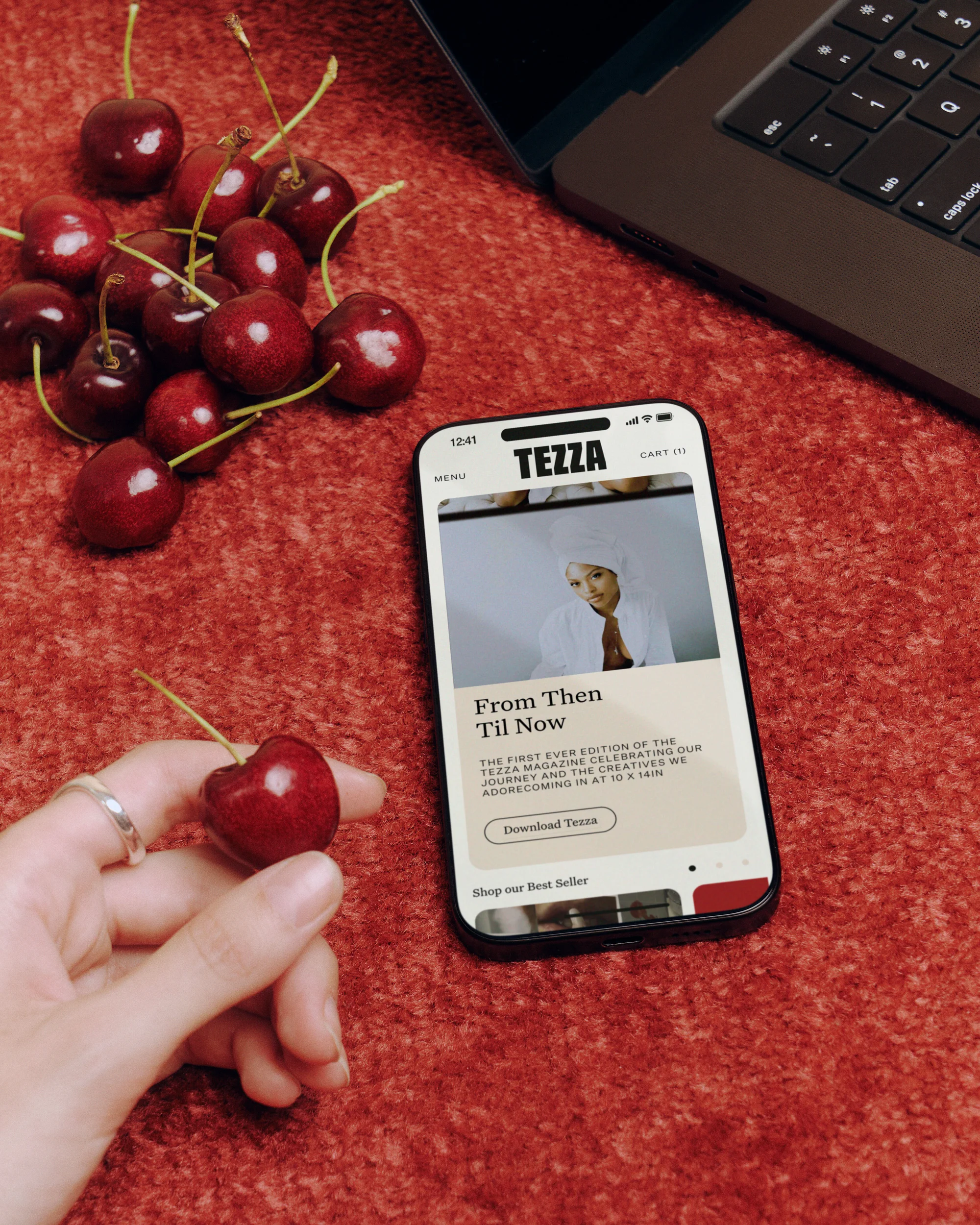 Close-up of a smartphone showing the Tezza app, with cherries, a hand holding a cherry, and a laptop in the background on a red surface.