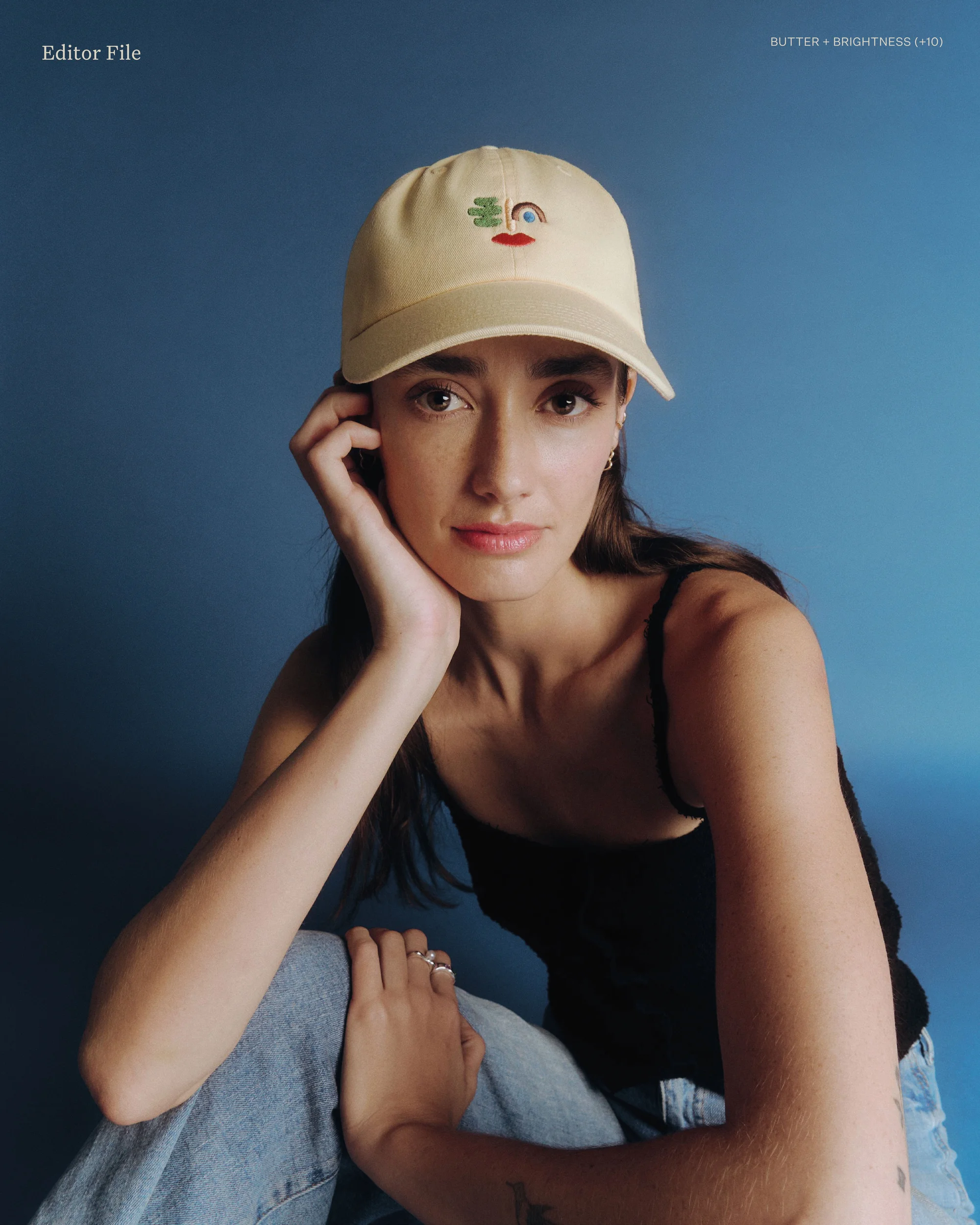 Portrait of a brunette woman wearing a baseball cap with a colorful design, seated in front of a solid blue backdrop with soft lighting.