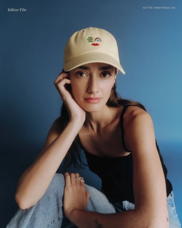 Portrait of a brunette woman wearing a baseball cap with a colorful design, seated in front of a solid blue backdrop with soft lighting.