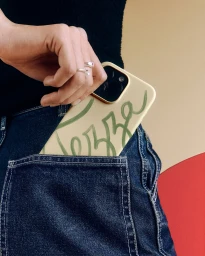 Close-up of a person holding a Tezza branded phone case tucked into the back pocket of dark blue denim jeans against a geometric background.