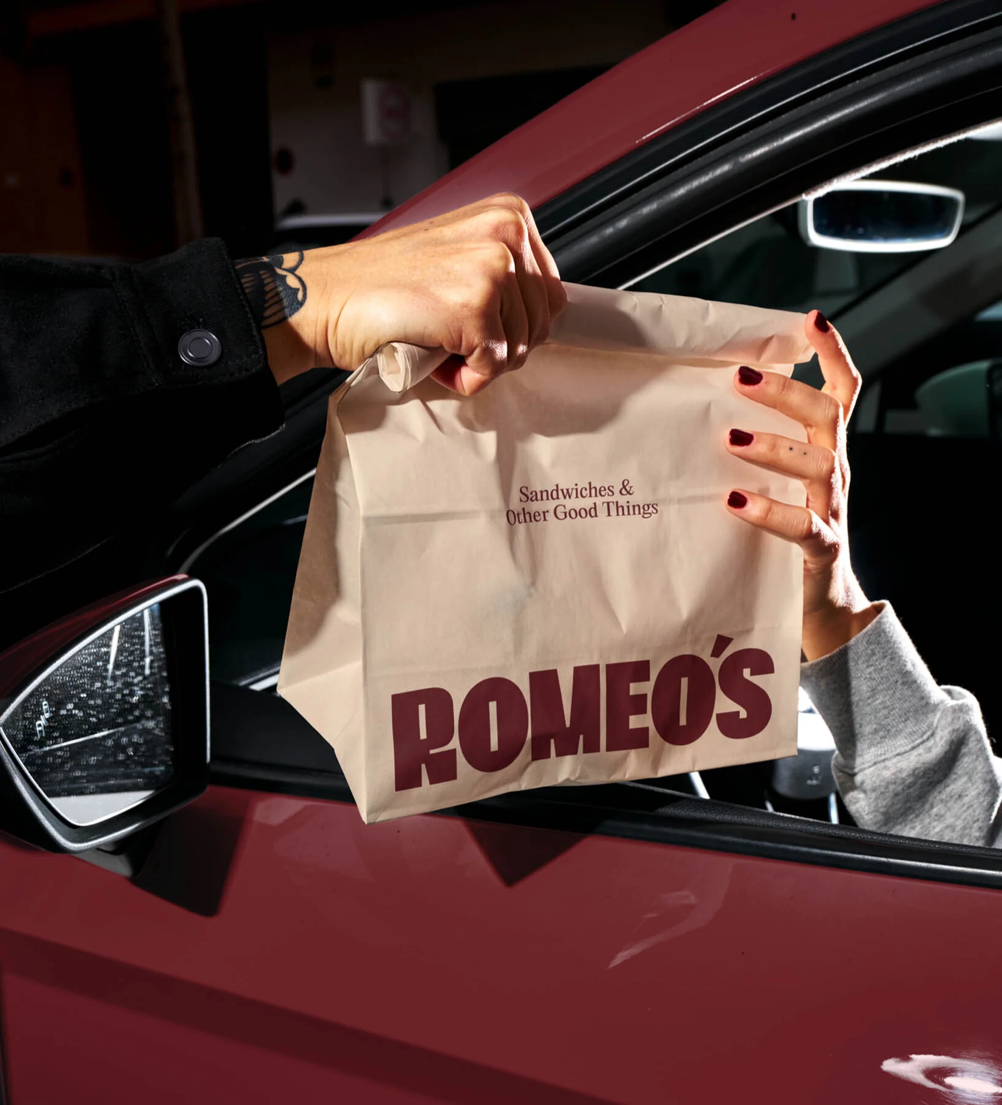 Hands exchange a takeout bag from a car window, with a dark red car and visible text on the bag, promoting a restaurant.