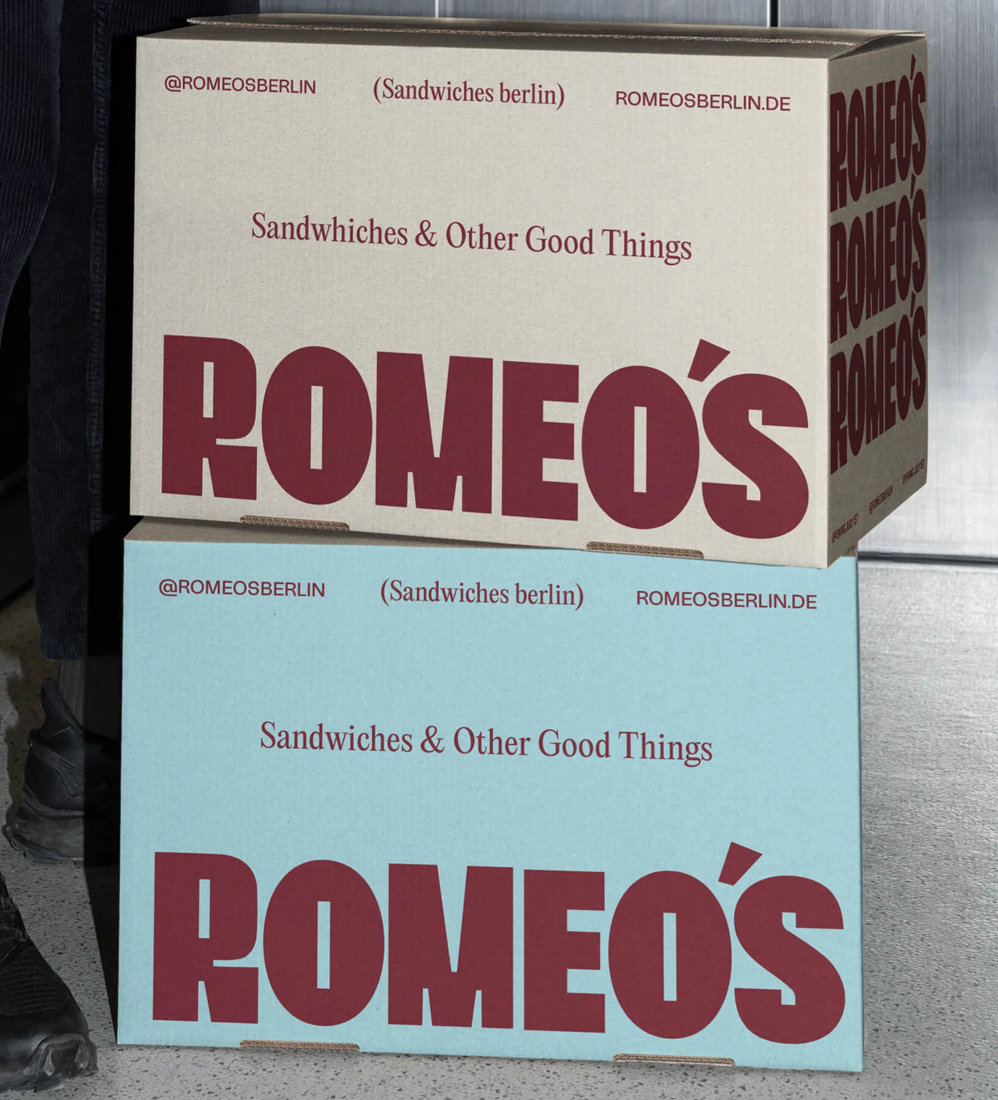 Two stacked cardboard boxes, one beige and one light blue, each labeled with the words 'Sandwiches & Other Good Things' and 'Romeo's'.