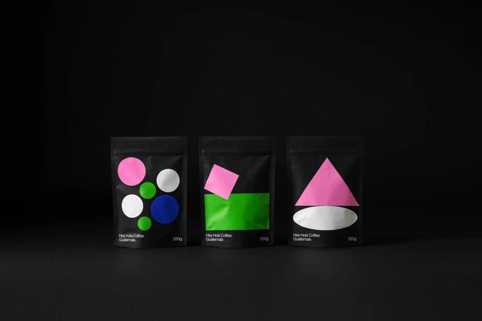 Three black coffee bags with pink, blue, green, and white geometric designs on a dark background, minimalist style, product photography.
