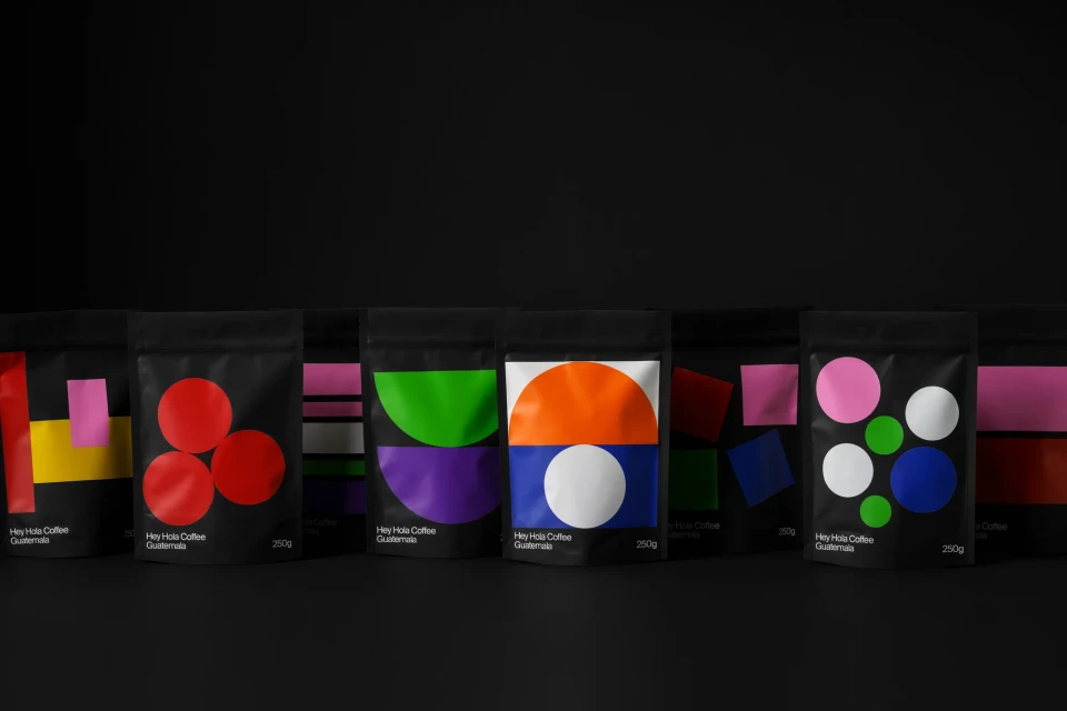 Several black pouches of Hey Hola Coffee Guatemala are arranged in a row against a dark background, each with colorful geometric designs.
