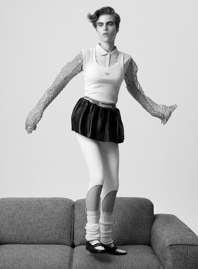 Monochrome studio shot of a woman in a knit top, black skirt, leggings, leg warmers, and Mary Jane shoes posing on a gray couch.