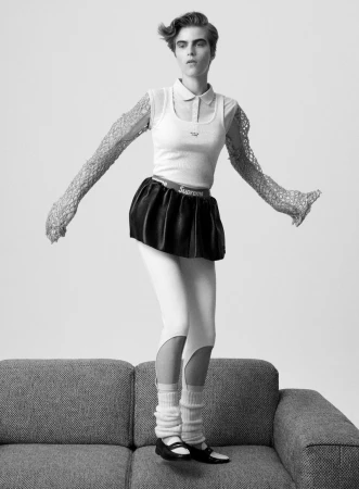 Monochrome studio shot of a woman in a knit top, black skirt, leggings, leg warmers, and Mary Jane shoes posing on a gray couch.