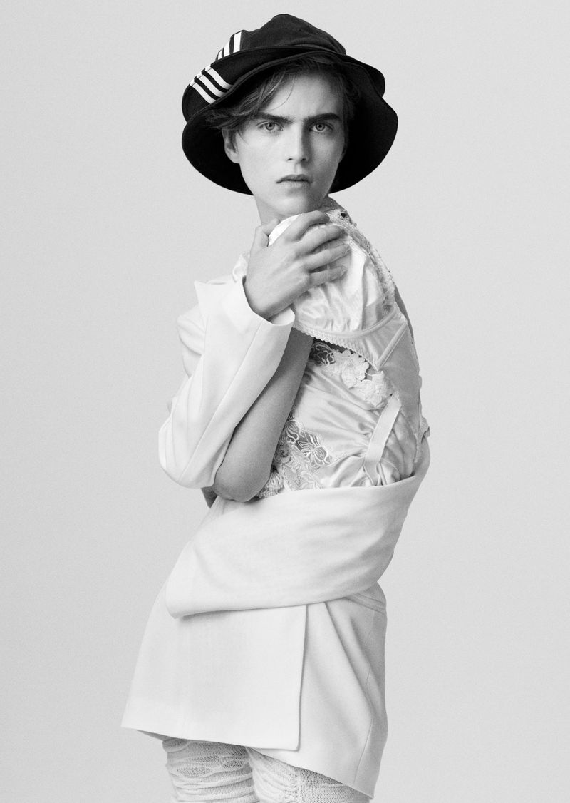 Black and white portrait of a person in a dark bucket hat with white stripes and a white suit jacket, standing in a photo studio.