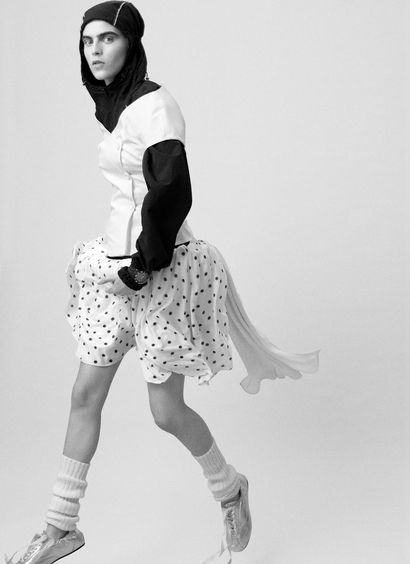 Black and white fashion shot of a person in a black head covering, white top, polka dot shorts, leg warmers and silver shoes.