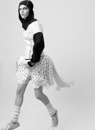 Black and white fashion shot of a person in a black head covering, white top, polka dot shorts, leg warmers and silver shoes.