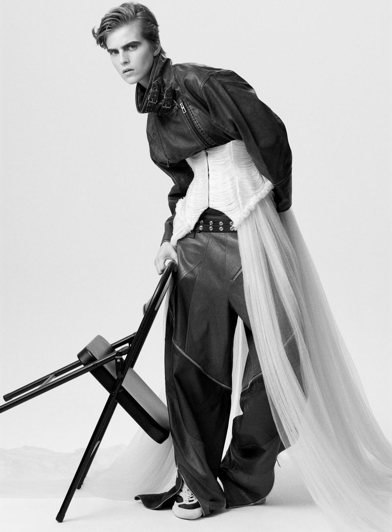Black and white fashion shot of a person wearing a leather jacket, corset, and tulle skirt, posed next to a folding chair in a studio.