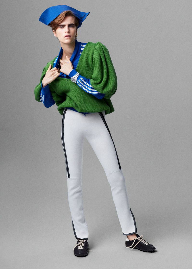 A person in a green sweatshirt and white pants poses in a studio against a gray background, wearing a blue tricorne hat.