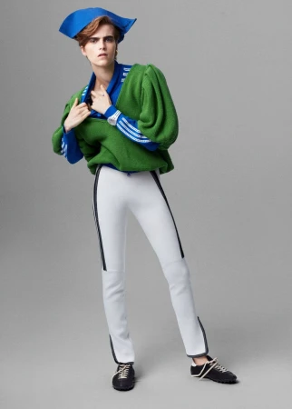A person in a green sweatshirt and white pants poses in a studio against a gray background, wearing a blue tricorne hat.