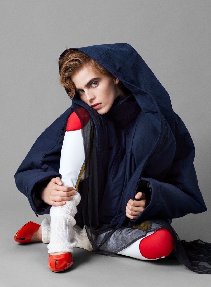 Fashion shot of a model in a hooded blue jacket with red shoes, posing in a seated position on a gray background in a studio.