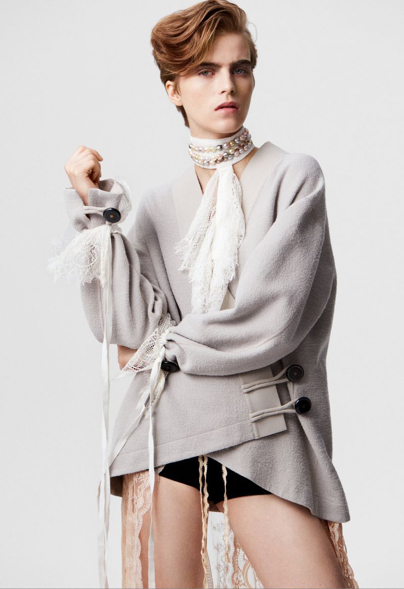 Adult female fashion model poses in a studio wearing a grey oversized sweater and statement necklace in a portrait-style shot.