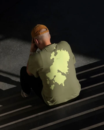 An adult sits on stairs wearing a tan hat and light-green t-shirt with a map graphic; shot from the back in shadow and natural light.