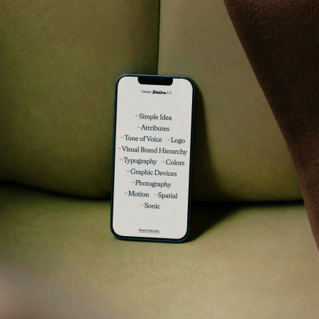 Mobile phone displaying design elements on a light green surface with a blurred beige fabric background. Branding and identity mockup.