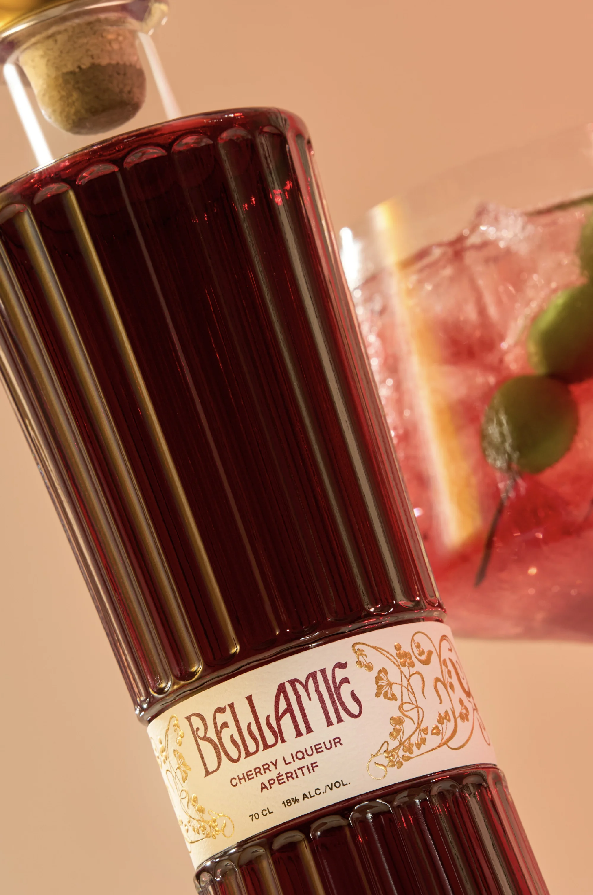 Ribbed bottle of Bellamie cherry liqueur with a cork stopper and an iced cocktail with fruit garnish in the background, indoors.