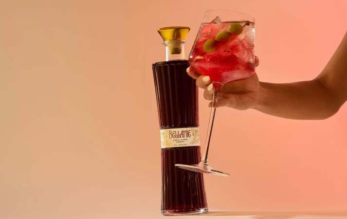A bottle of cherry liqueur and a wine glass of a cherry liqueur cocktail are held in front of a peach gradient background.
