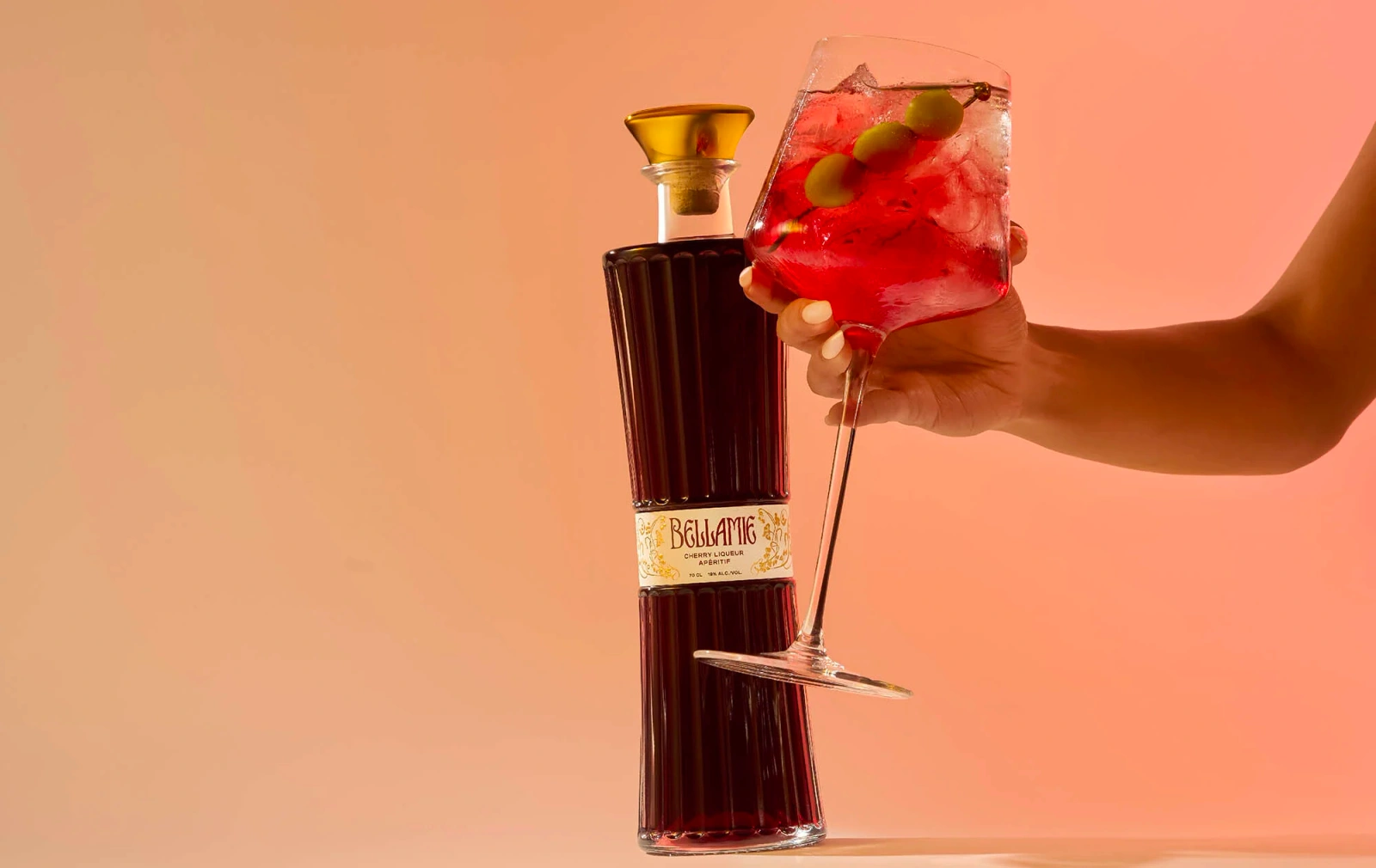 A person holding a large red cocktail glass with olives next to a tall ribbed Bellamie liquor bottle on a warm gradient background.
