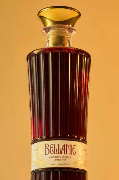Tall fluted glass bottle of dark cherry liqueur with a gold stopper against a warm golden background