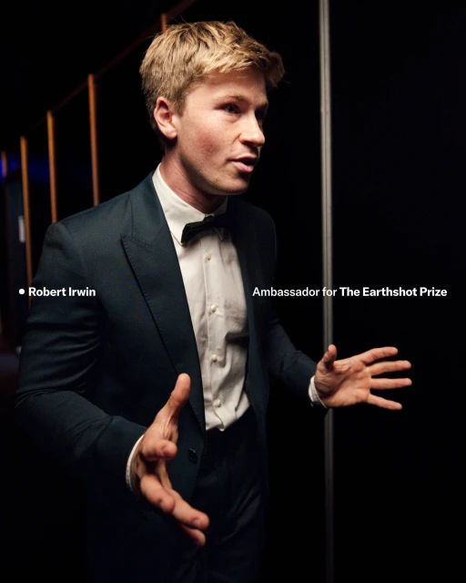 Robert Irwin, environmental advocate, stands in a suit and bowtie against a black backdrop with side lighting, promoting The Earthshot Prize.