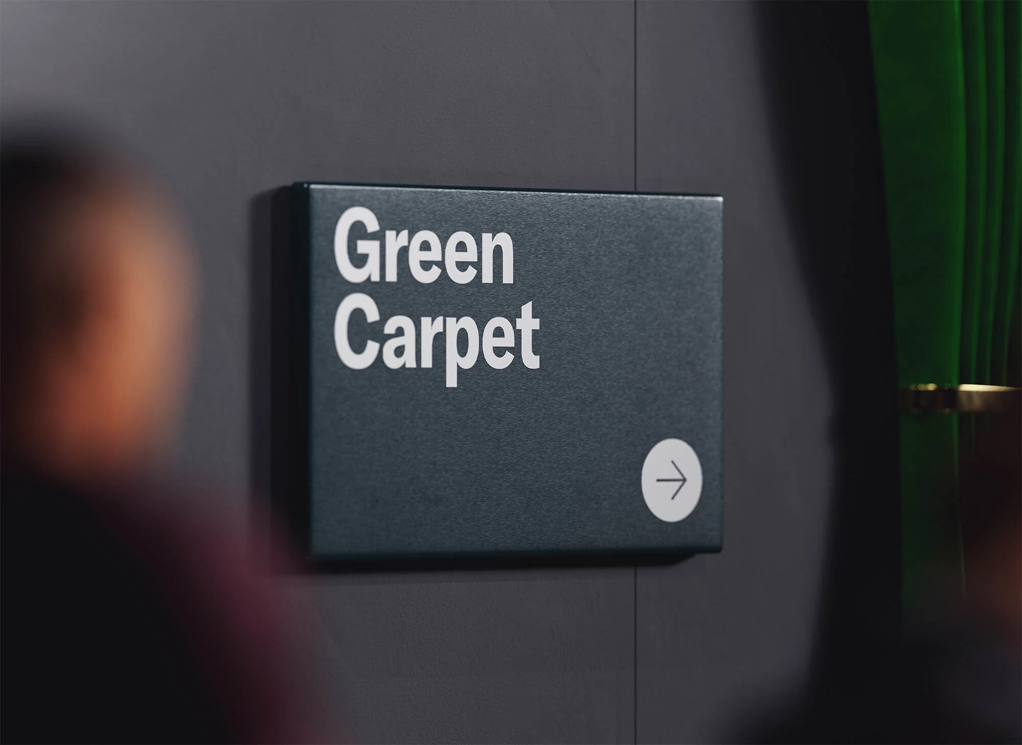 Sign reading 'Green Carpet' with a right arrow, mounted on a gray wall, with a blurred person and a green curtain in the background.