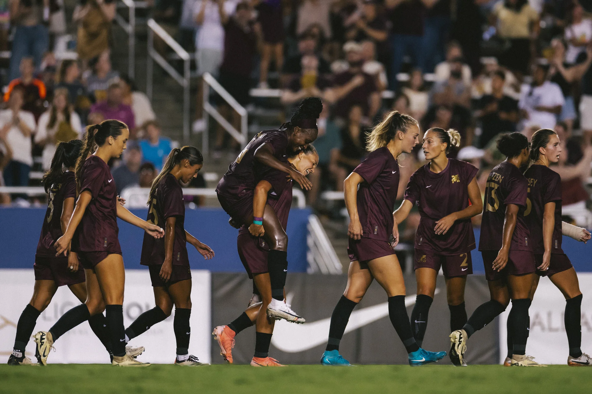 Female soccer players celebrating on the field in maroon uniforms. One athlete gives another a piggyback ride during a match.
