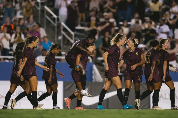Female soccer players celebrating on the field in maroon uniforms. One athlete gives another a piggyback ride during a match.