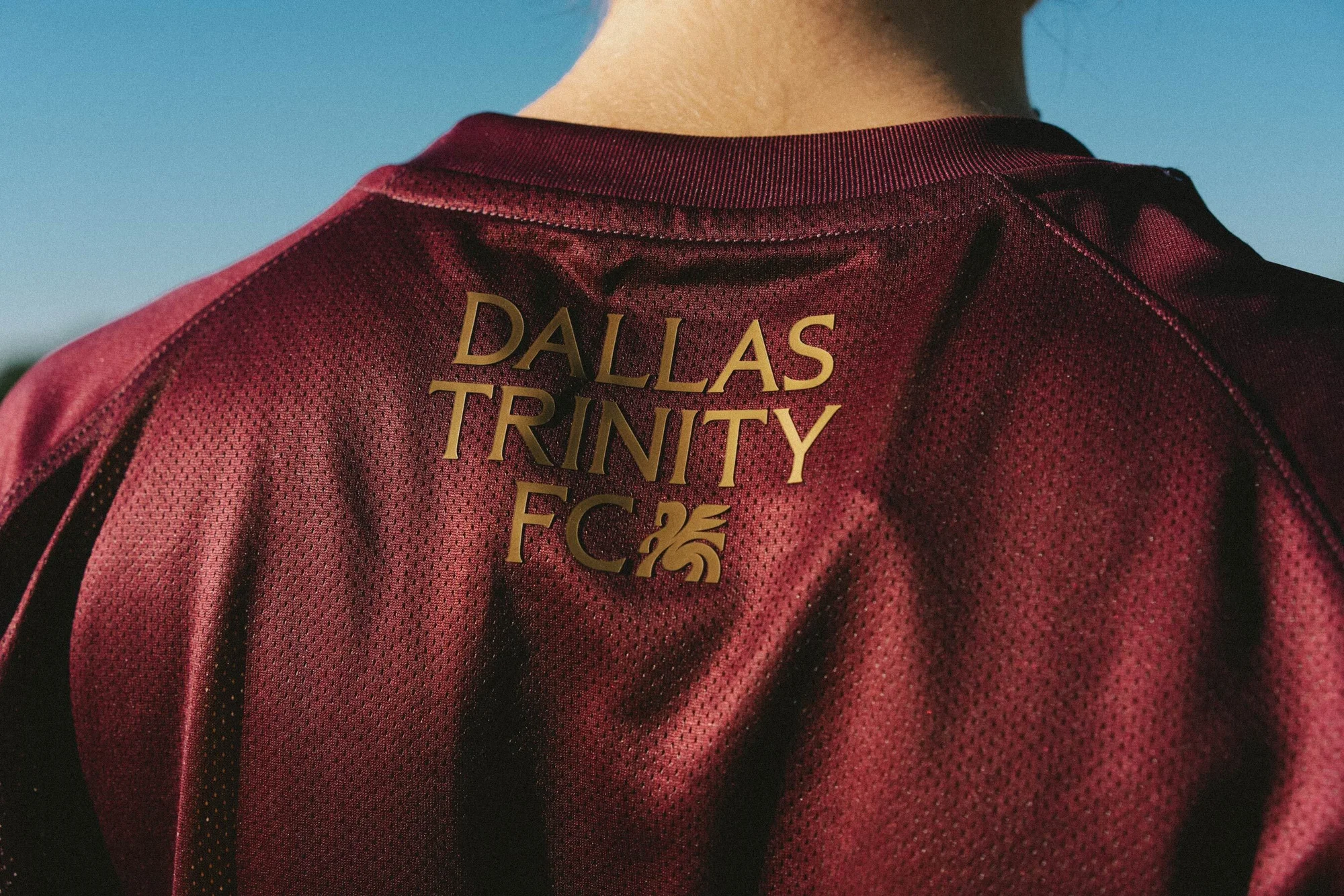 Close-up of a person wearing a maroon soccer jersey with 'Dallas Trinity FC' printed in gold, against a blue sky background.