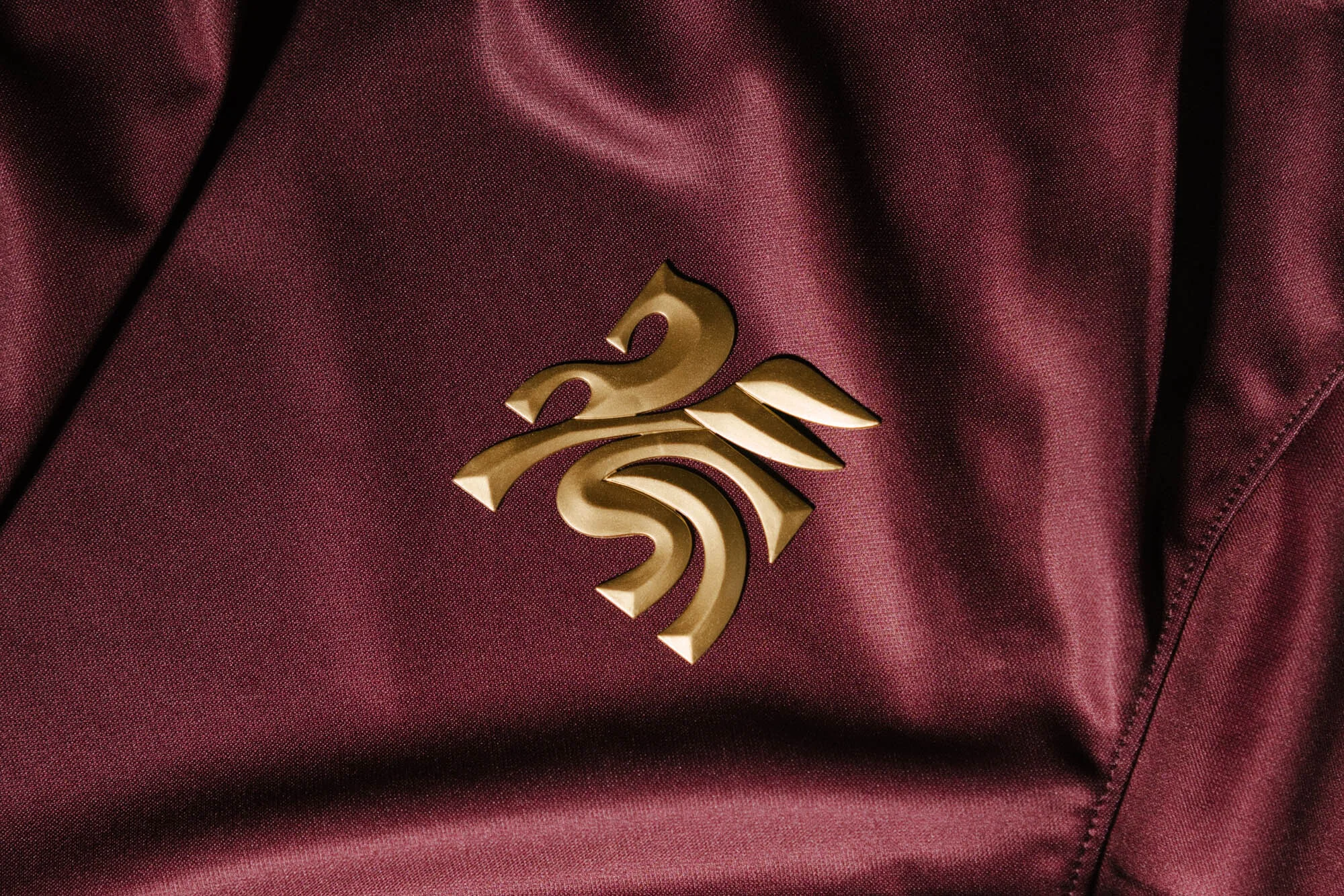 Close-up of a burgundy satin fabric featuring an ornate golden logo. The material has a shiny finish with folds and a tailored seam.