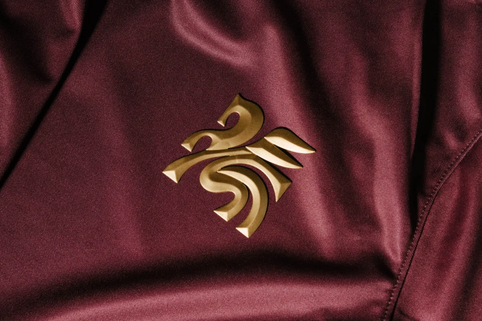 Close-up of a burgundy satin fabric featuring an ornate golden logo. The material has a shiny finish with folds and a tailored seam.