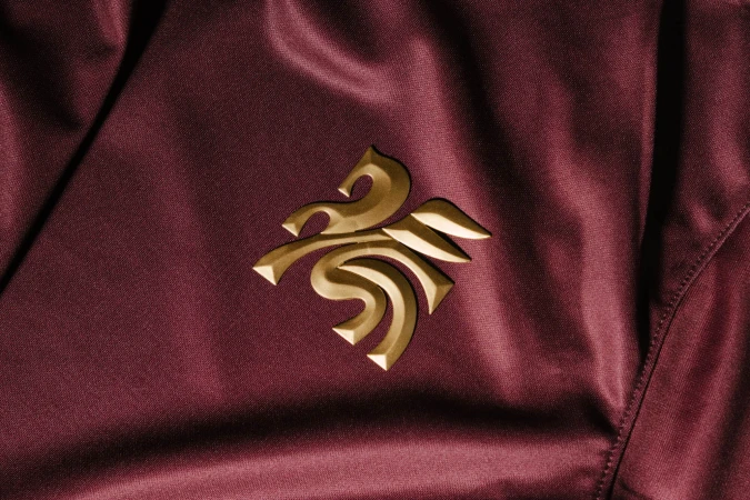 Close-up of a burgundy satin fabric featuring an ornate golden logo. The material has a shiny finish with folds and a tailored seam.
