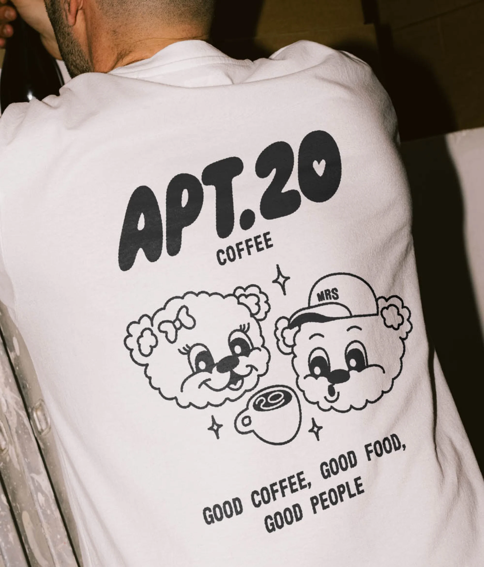 Person wearing a white t-shirt with a cartoon graphic of two bear faces, a coffee cup and the text APT.20 COFFEE printed on it.