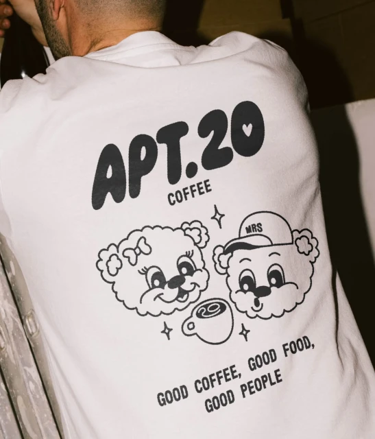 Person wearing a white t-shirt with a cartoon graphic of two bear faces, a coffee cup and the text APT.20 COFFEE printed on it.