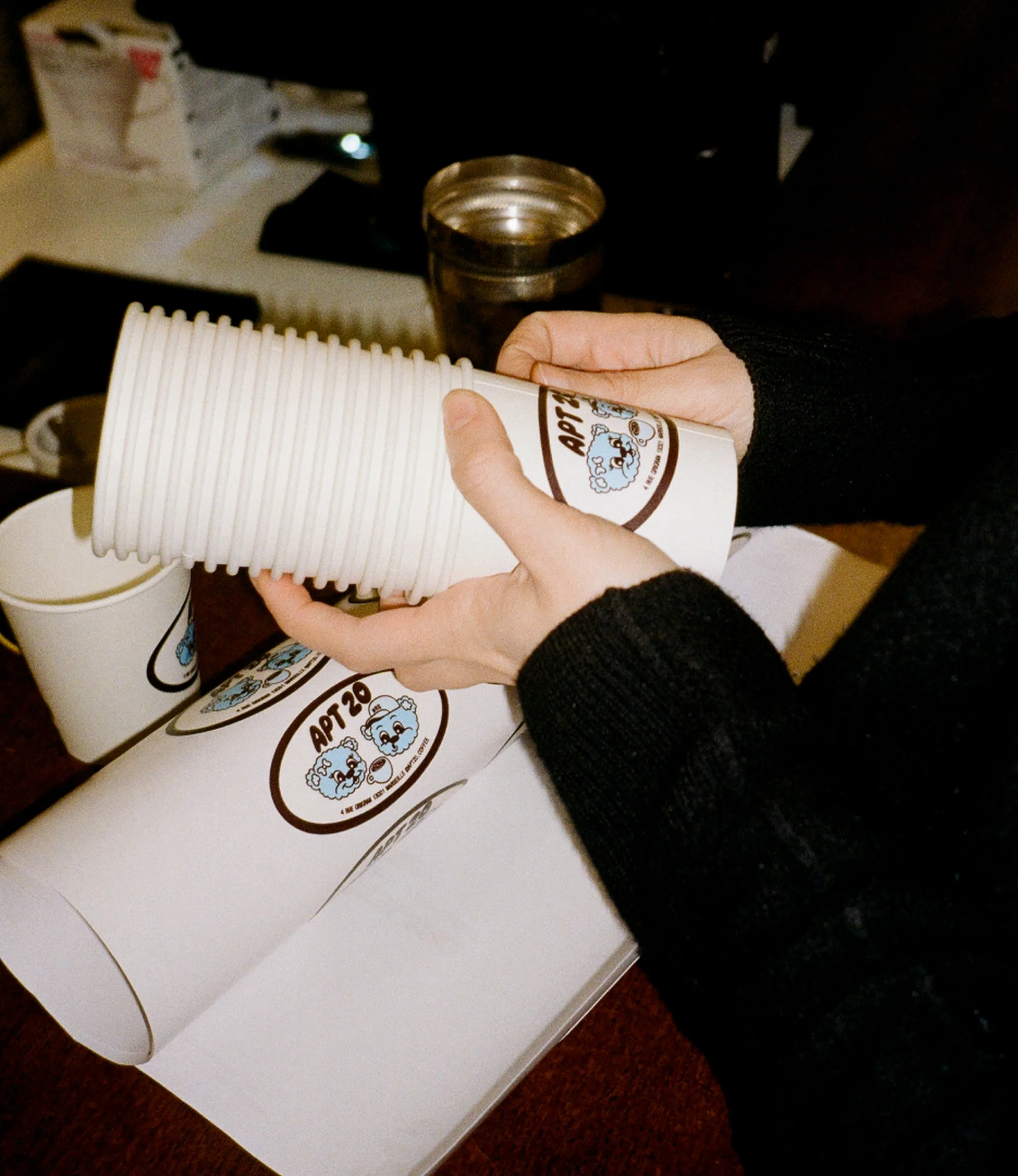 Close-up of hands holding a stack of white paper cups with a blue bear logo, alongside printed logo sheets on a dark wooden surface.