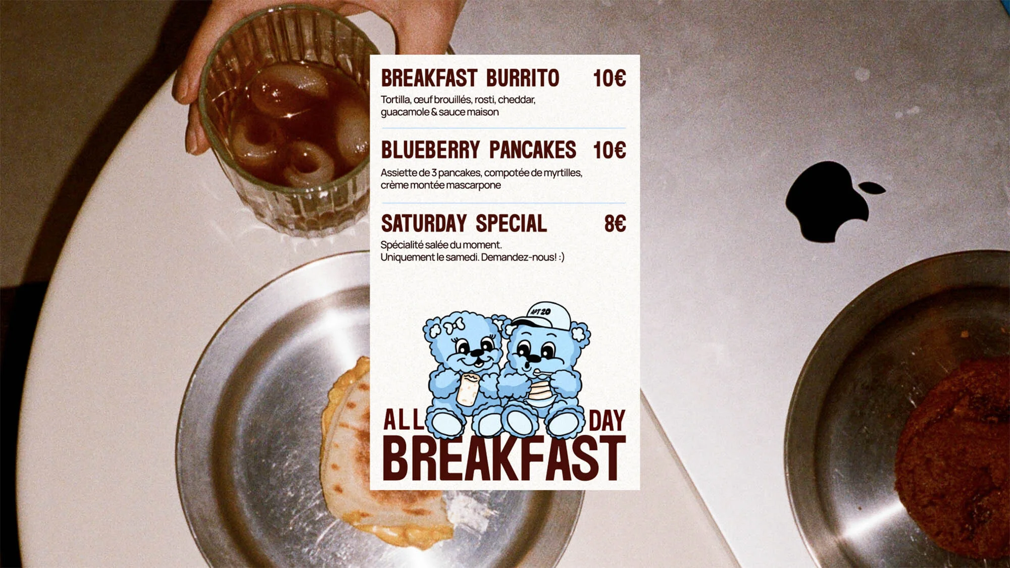 Overhead shot of breakfast foods with a menu featuring breakfast burrito and blueberry pancakes next to an Apple laptop.
