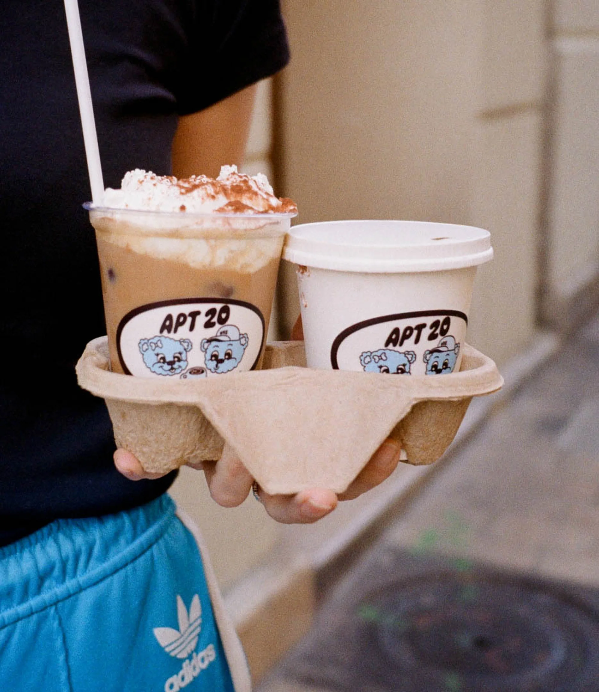 Iced coffee and covered coffee beverage sit in a cardboard carrier held by a person wearing Adidas shorts, capturing a casual, cafe moment.