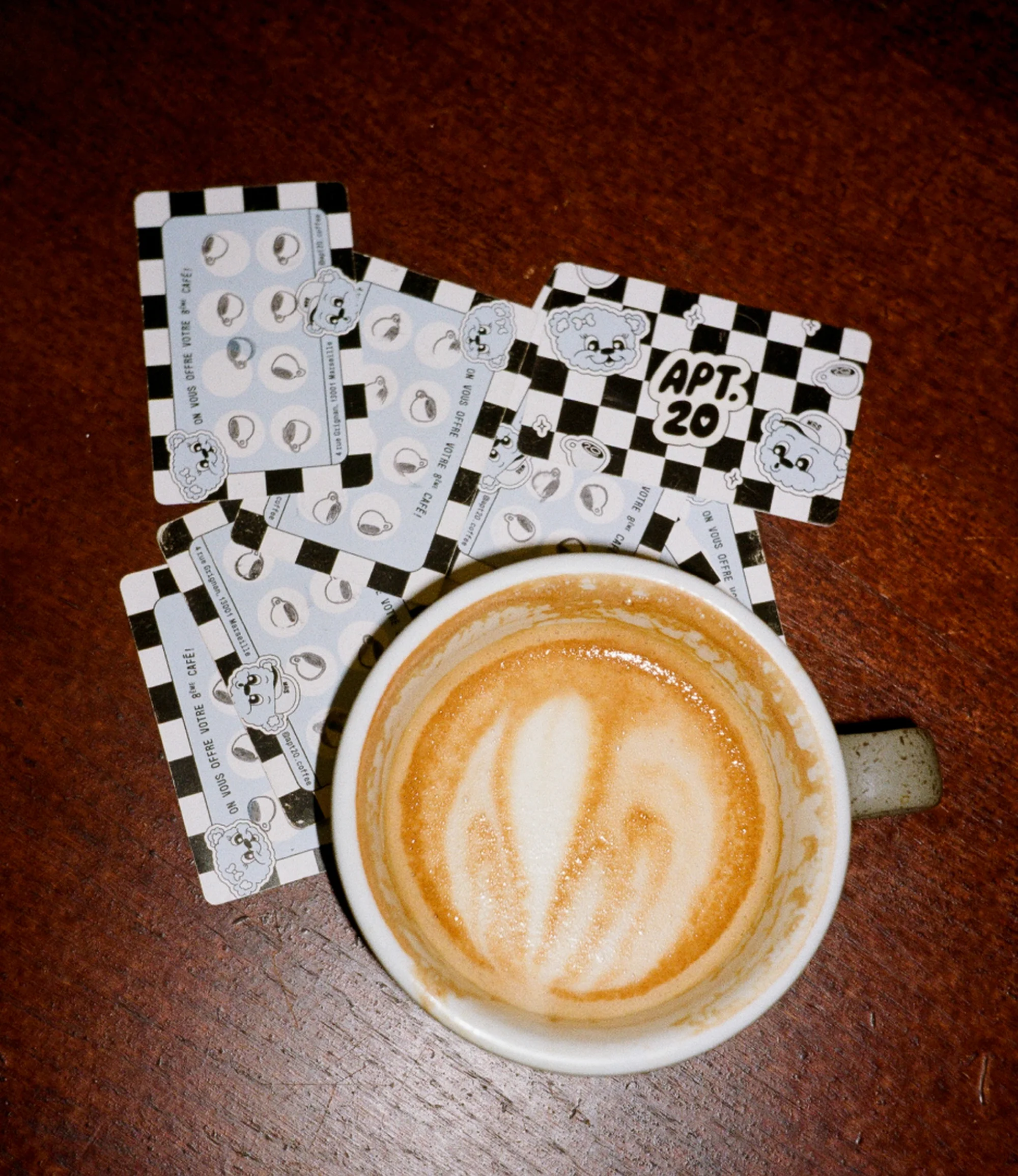 Elevated view of coffee cup with latte art on a wood table, along with several APT 20 cafe cards with cartoon illustrations.