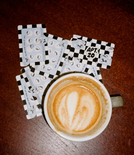 Elevated view of coffee cup with latte art on a wood table, along with several APT 20 cafe cards with cartoon illustrations.