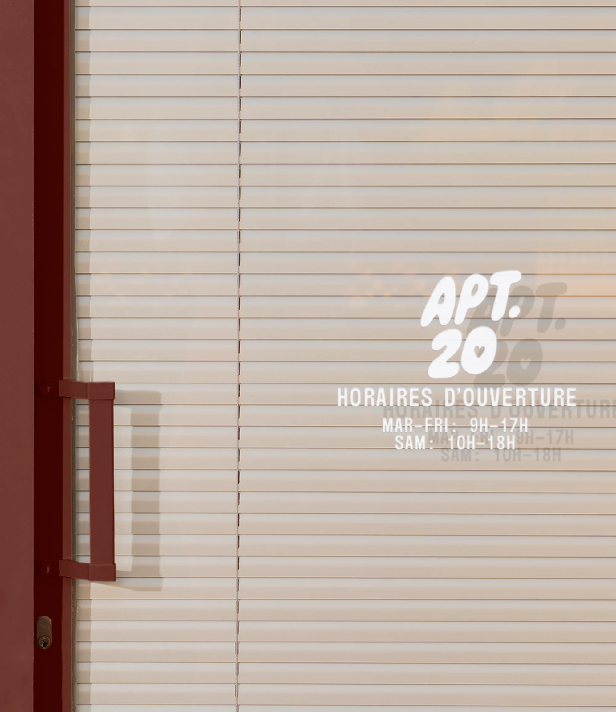 Close-up of a closed storefront door with blinds, featuring a small opening hours sign and the name "APT. 20" in white lettering.