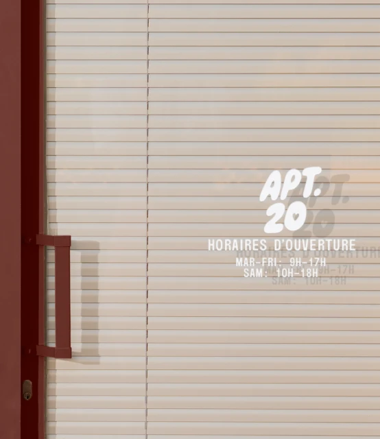 Close-up of a closed storefront door with blinds, featuring a small opening hours sign and the name "APT. 20" in white lettering.