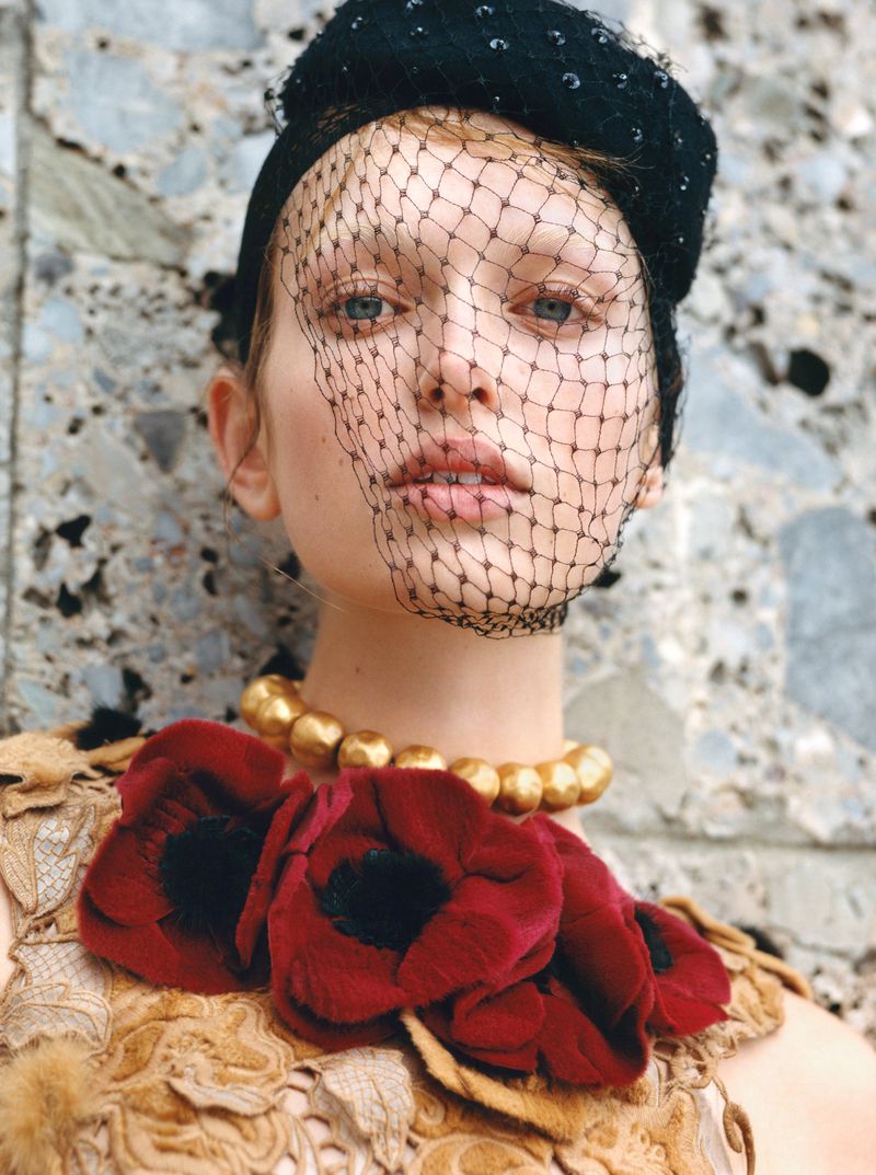 Portrait of a woman in a black hat with a net veil, wearing a gold beaded necklace and a floral-embellished top, against a stone wall.